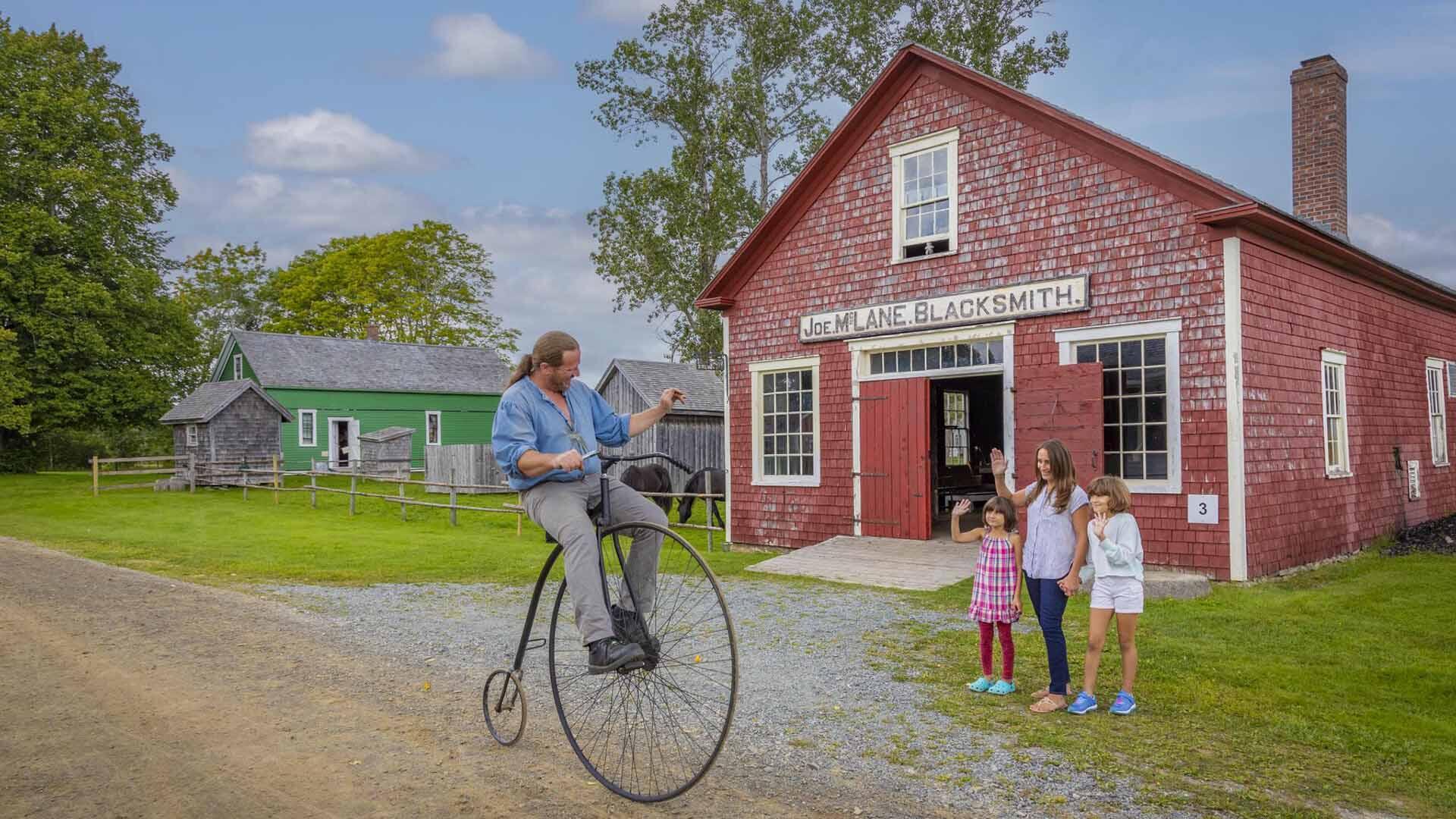 Man riding an antique bike past waving children in Sherbrooke Village, a living history museum, in Sherbrooke, Nova Scotia