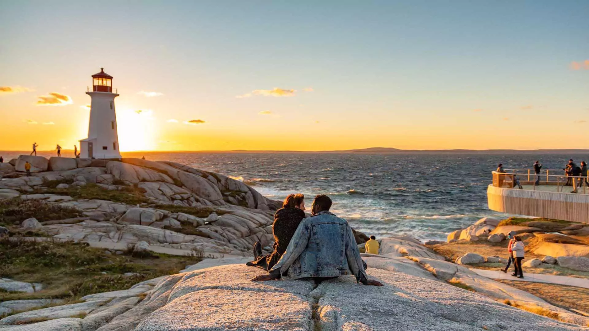 A couple sits on granite rock next to the viewing platform taking in the setting sun over the Atlantic Ocean behind the Peggy's Cove Lighthouse in Peggy's Cove, Nova Scotia.