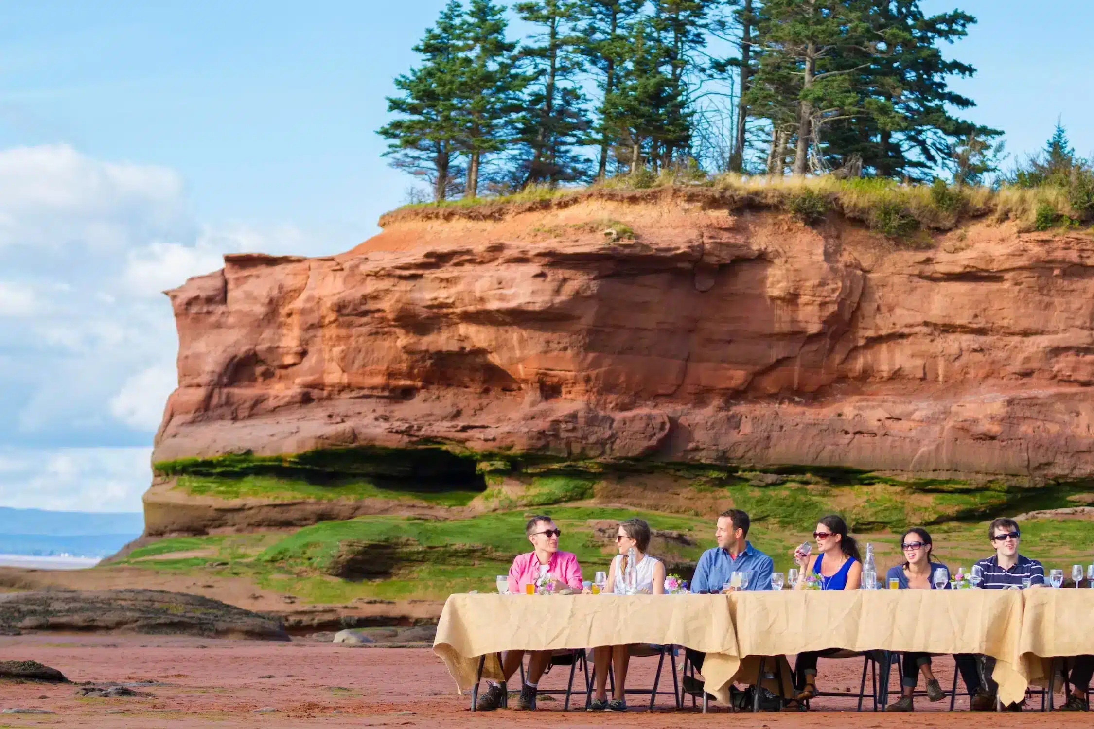 a group of people sitting at a long table having food on the beach