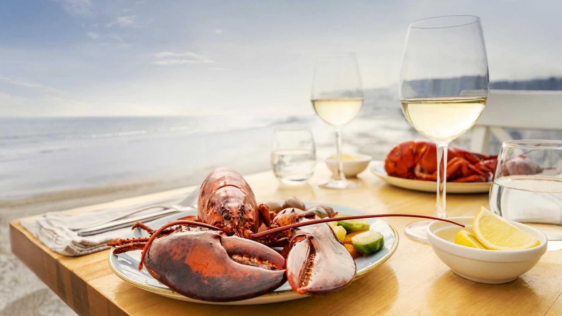 A plated boiled lobster dinner with seasonal summertime vegetables and Tidal Bay white wine on an outside table on the deck overlooking Summerville Beach on a sunny summer afternoon at The Quarterdeck Beachside Villas and Grill near Liverpool, Nova Scotia